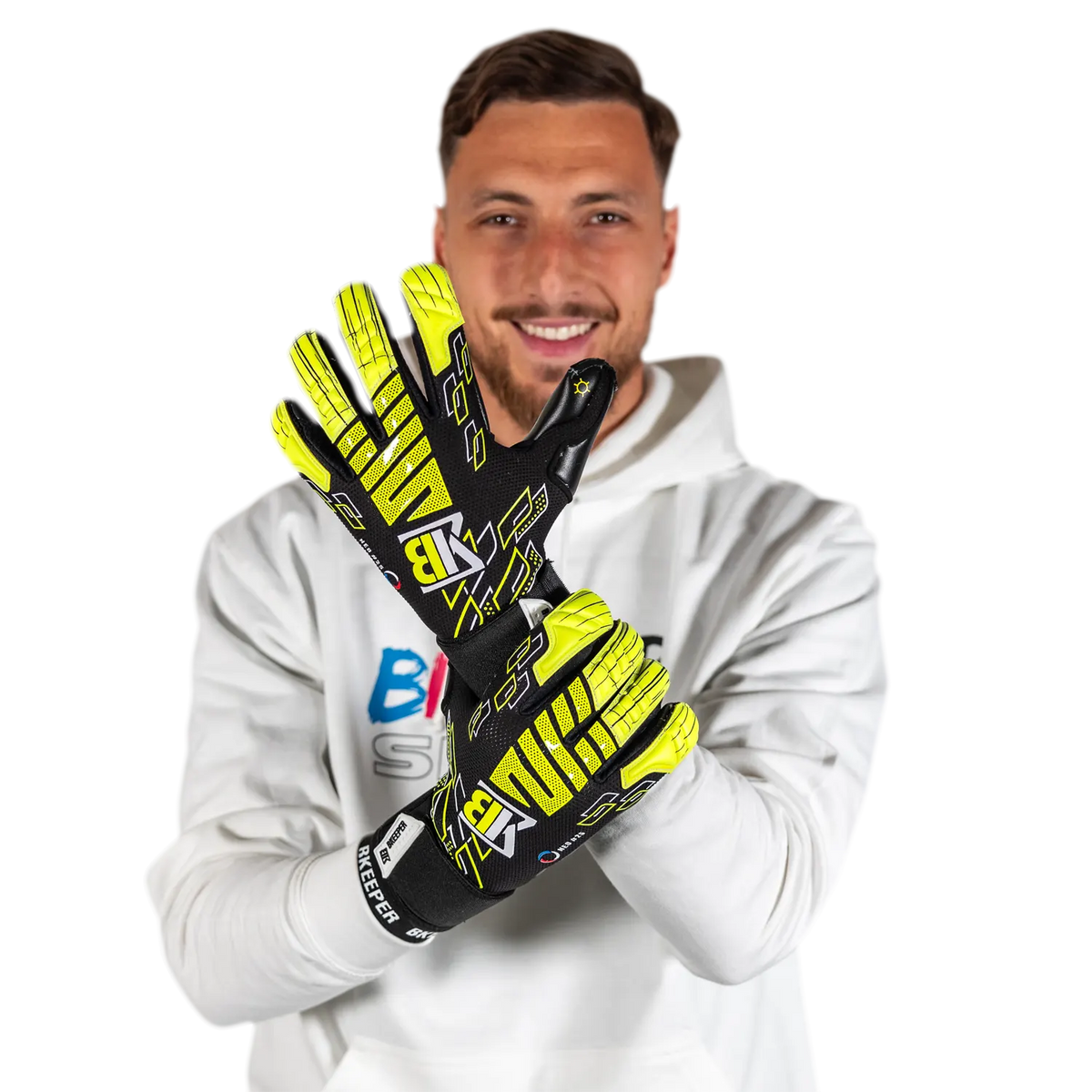 A man wearing a white hoodie holds up a pair of black and neon yellow goalkeeper gloves against a solid white background.