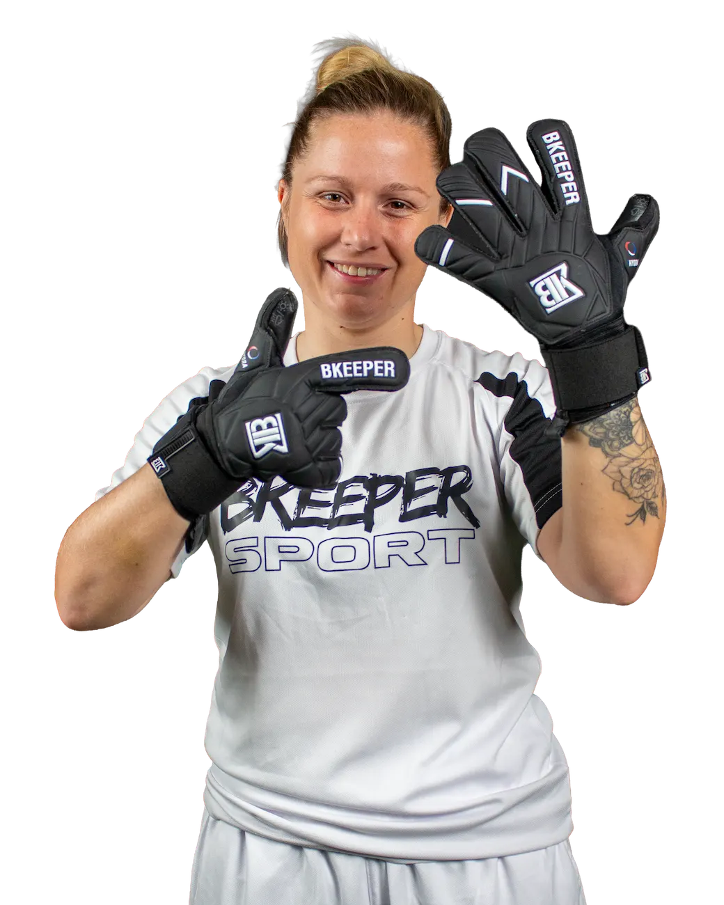 A smiling woman wearing a white Bkeeper Sport t-shirt and light gray shorts, holding up a pair of black goalkeeper gloves with white branding on a solid white background.